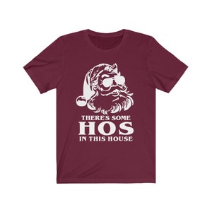 Pu&ograve; includere: T-shirt bordeaux con grafica bianca di Babbo Natale con occhiali da sole e cappello da Babbo Natale. Il testo "THERE'S SOME HOS IN THIS HOUSE" &egrave; stampato in bianco sotto.