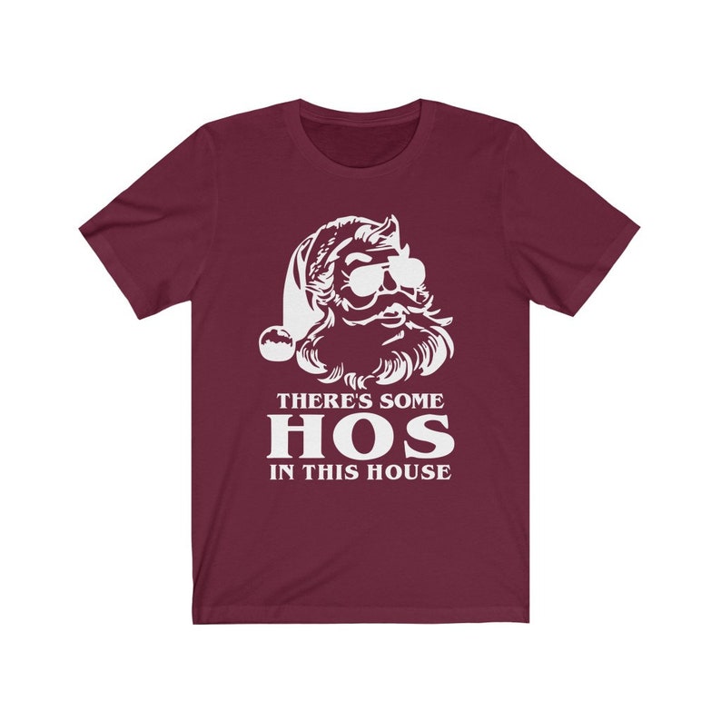 Pu&ograve; includere: T-shirt bordeaux con grafica bianca di Babbo Natale con occhiali da sole e cappello da Babbo Natale. Il testo "THERE'S SOME HOS IN THIS HOUSE" &egrave; stampato in bianco sotto.