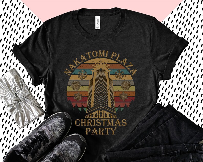 May include: Black t-shirt with a retro design featuring the text "NAKATOMI PLAZA CHRISTMAS PARTY." The design includes a skyscraper, snowflakes, and a sunset-style background. The shirt is paired with black jeans and sneakers.