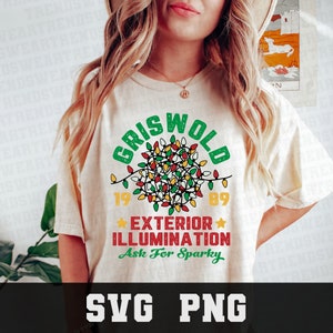 May include: Cream-colored t-shirt with a festive design. The shirt features the word "GRISWOLD" in green above a cluster of colorful Christmas lights. Below the lights are the numbers "1989" and the words "EXTERIOR ILLUMINATION" in red. The phrase "Ask For Sparky" is in green.