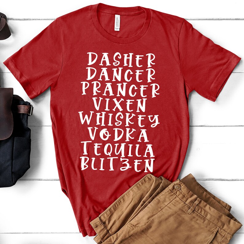 May include: A red t-shirt with white text listing the names "Dasher, Dancer, Prancer, Vixen, Whiskey, Vodka, Tequila, and Blitzen". The shirt is displayed on a white wooden surface next to brown trousers and a black bag.