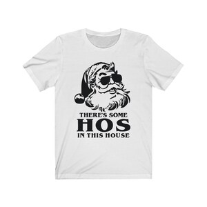 Pu&ograve; includere: T-shirt bianca con grafica nera di Babbo Natale con occhiali da sole. Il testo "THERE'S SOME HOS IN THIS HOUSE" &egrave; stampato sotto. La maglietta ha maniche corte e girocollo, adatta per un look casual.