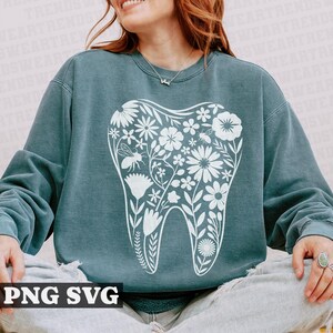 May include: A teal blue sweatshirt with a white floral tooth design. The tooth is surrounded by flowers and leaves. The sweatshirt is a comfortable and stylish option for anyone who loves dental care.