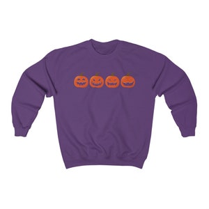 K&ouml;nnte beinhalten: Ein lila Sweatshirt mit vier orangefarbenen K&uuml;rbissen mit geschnitzten Gesichtern. Die K&uuml;rbisse haben gr&uuml;ne Stiele und sind horizontal angeordnet. Das Sweatshirt besteht aus einem weichen Material.