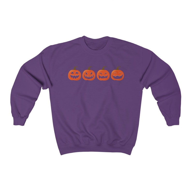 K&ouml;nnte beinhalten: Ein lila Sweatshirt mit vier orangefarbenen K&uuml;rbissen mit geschnitzten Gesichtern. Die K&uuml;rbisse haben gr&uuml;ne Stiele und sind horizontal angeordnet. Das Sweatshirt besteht aus einem weichen Material.