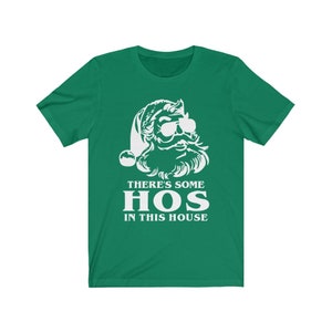 Pu&ograve; includere: T-shirt verde con grafica bianca di Babbo Natale con occhiali da sole e barba. Il testo "THERE'S SOME HOS IN THIS HOUSE" &egrave; stampato sotto. Un design divertente e festivo.