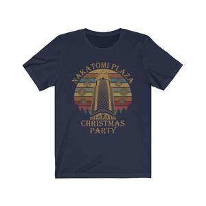 May include: Navy blue t-shirt featuring a retro design with the text "NAKATOMI PLAZA CHRISTMAS PARTY." The graphic includes a tall building, a sunset, and snowflakes. The shirt is made of a soft, comfortable material.