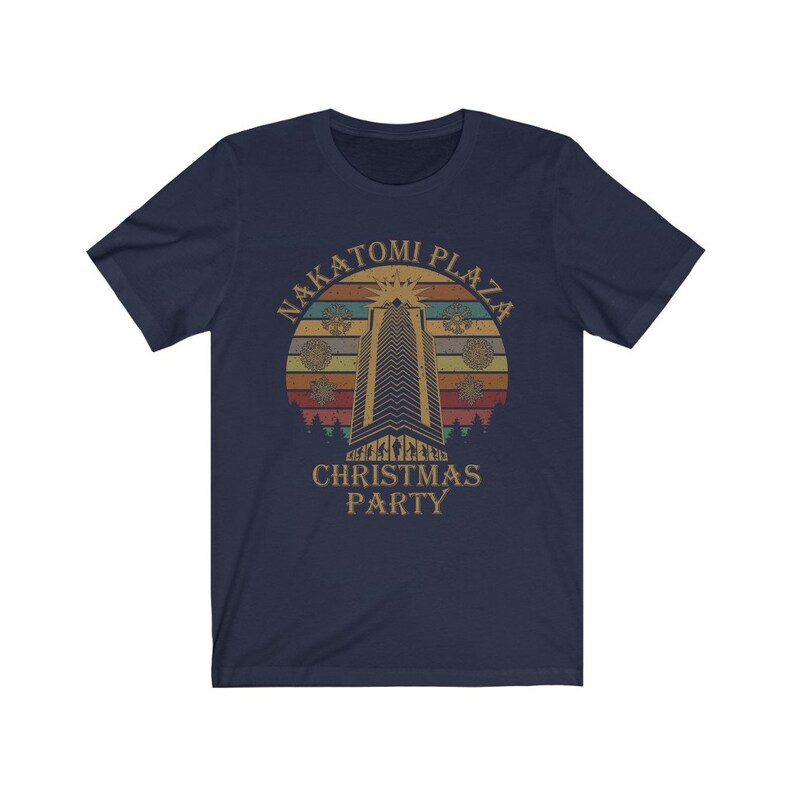 May include: Navy blue t-shirt featuring a retro design with the text "NAKATOMI PLAZA CHRISTMAS PARTY." The graphic includes a tall building, a sunset, and snowflakes. The shirt is made of a soft, comfortable material.