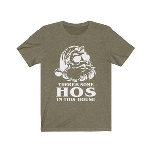 Pu&ograve; includere: T-shirt verde oliva con grafica bianca di Babbo Natale con occhiali da sole. Il testo "THERE'S SOME HOS IN THIS HOUSE" &egrave; stampato sotto l'illustrazione di Babbo Natale in un carattere grassetto senza grazie. Un design umoristico e festivo.