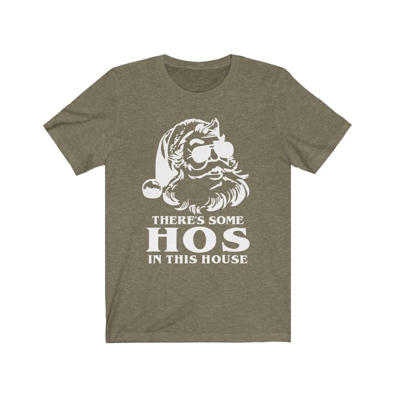 Pu&ograve; includere: T-shirt verde oliva con grafica bianca di Babbo Natale con occhiali da sole. Il testo "THERE'S SOME HOS IN THIS HOUSE" &egrave; stampato sotto l'illustrazione di Babbo Natale in un carattere grassetto senza grazie. Un design umoristico e festivo.