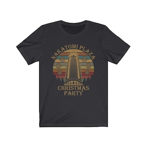 May include: Dark gray t-shirt featuring a retro design with the text "NAKATOMI PLAZA CHRISTMAS PARTY." The graphic includes a tall building, snowflakes, and a sunset-style background in shades of orange, yellow, and brown.