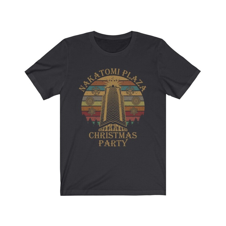 May include: Dark gray t-shirt featuring a retro design with the text "NAKATOMI PLAZA CHRISTMAS PARTY." The graphic includes a tall building, snowflakes, and a sunset-style background in shades of orange, yellow, and brown.