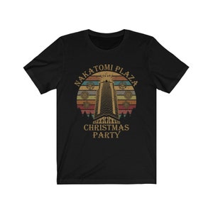 May include: Black t-shirt featuring a retro design with the text "Nakatomi Plaza Christmas Party." The design includes a tall building, a sunset, and decorative elements, evoking a vintage aesthetic.