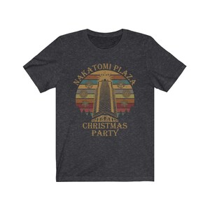 May include: Dark gray t-shirt featuring a retro design with the text "NAKATOMI PLAZA CHRISTMAS PARTY." The graphic includes a tall building, a sunburst, and snowflakes, set against a sunset-colored background. The shirt is made of a soft material.