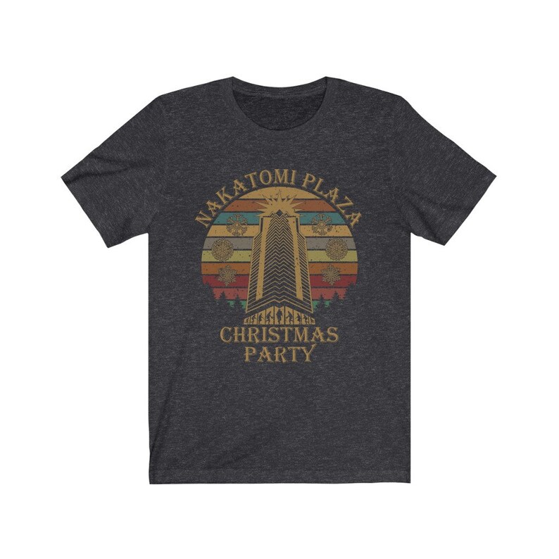 May include: Dark gray t-shirt featuring a retro design with the text "NAKATOMI PLAZA CHRISTMAS PARTY." The graphic includes a tall building, a sunburst, and snowflakes, set against a sunset-colored background. The shirt is made of a soft material.
