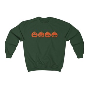 K&ouml;nnte beinhalten: Ein dunkelgr&uuml;nes Sweatshirt mit vier orangefarbenen K&uuml;rbisgesichtern mit unterschiedlichen Schnitzereien. Die K&uuml;rbisse sind horizontal angeordnet. Das Sweatshirt hat einen Rundhalsausschnitt und lange &Auml;rmel, ideal f&uuml;r Halloween.