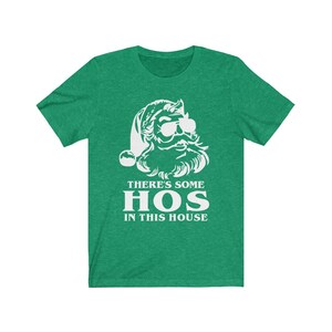 Pu&ograve; includere: T-shirt verde con grafica bianca di Babbo Natale con occhiali da sole e barba. Il testo "THERE'S SOME HOS IN THIS HOUSE" &egrave; stampato in bianco sotto. La maglietta &egrave; girocollo.