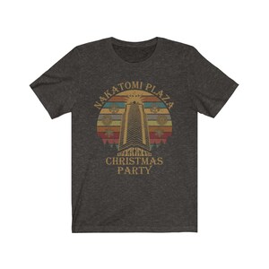 May include: Dark heather gray t-shirt featuring a retro design with the text "Nakatomi Plaza Christmas Party". The graphic includes a tall building, snowflakes, and a sunset-style background.