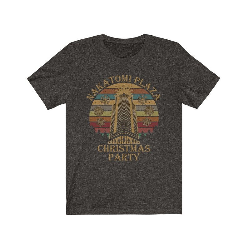 May include: Dark heather gray t-shirt featuring a retro design with the text "Nakatomi Plaza Christmas Party". The graphic includes a tall building, snowflakes, and a sunset-style background.