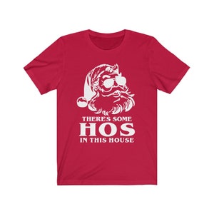 Pu&ograve; includere: T-shirt rossa con grafica bianca di Babbo Natale con occhiali da sole. Il testo "THERE'S SOME HOS IN THIS HOUSE" &egrave; stampato sotto la grafica di Babbo Natale in lettere bianche.