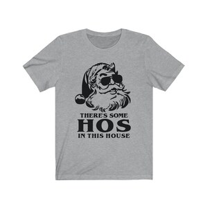 Pu&ograve; includere: T-shirt grigia con grafica nera di Babbo Natale con occhiali da sole. Il testo "THERE'S SOME HOS IN THIS HOUSE" &egrave; stampato sotto. La maglietta ha maniche corte e girocollo. Il design &egrave; umoristico.