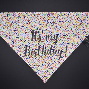 May include: A white dog bandana with a colorful sprinkle pattern and the text "It's my Birthday!" in black script. The bandana is triangular and has a small black tag attached to the side.