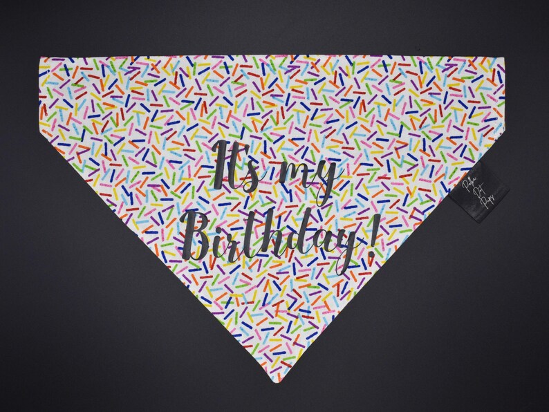 May include: A white dog bandana with a colorful sprinkle pattern and the text "It's my Birthday!" in black script. The bandana is triangular and has a small black tag attached to the side.