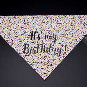 May include: White dog bandana with a colorful sprinkle pattern and the text "It's my Birthday!" in black. A small black tag is attached to the side. The bandana is triangular in shape.