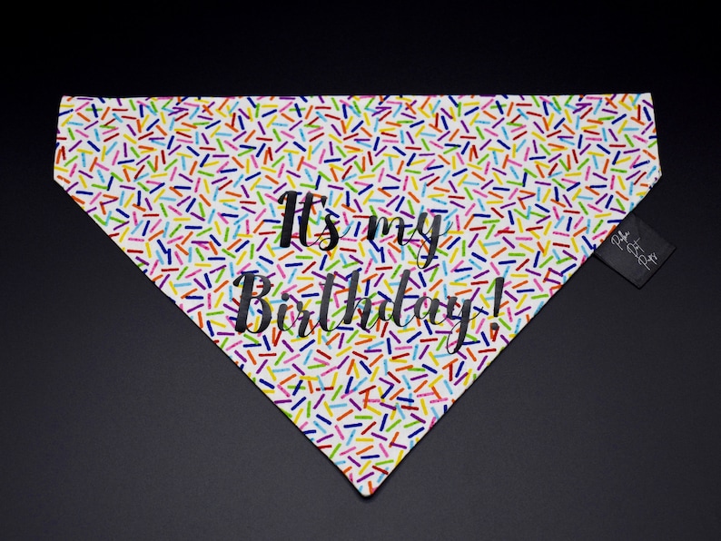 May include: White dog bandana with a colorful sprinkle pattern and the text "It's my Birthday!" in black. A small black tag is attached to the side. The bandana is triangular in shape.