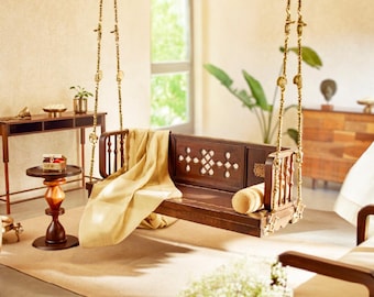May include: A dark brown wooden indoor swing suspended by ornate gold-toned metal chains. The swing features a backrest with a geometric cutout pattern and is draped with a light yellow fabric throw. A cylindrical bolster pillow rests on the seat.
