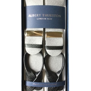 May include: A pair of gray suspenders with black leather ends and gold metal clips. The suspenders are in a blue box with the text "ALBERT THURSTON LONDON 1820" and "Handmade in England".