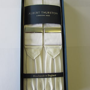 Peut inclure: Bretelles blanches avec des accents en cuir et des clips en métal doré. Les bretelles sont emballées dans une boîte noire avec le texte "ALBERT THURSTON LONDON 1820" et "Fabriqué en Angleterre".