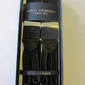 Peut inclure: Bretelles noires avec des attaches argentées, présentées dans une boîte bleu foncé. Les bretelles comportent des détails en cuir et le texte "ALBERT THURSTON LONDON 1820" et "Handmade in England".