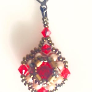 Może przedstawiać: Ręcznie robiony kolczyk w kształcie łzy. Posiada centralny czerwony kamień szlachetny, otoczony mniejszymi czerwonymi i złotymi koralikami. Kolczyk ma czarne zapięcie.