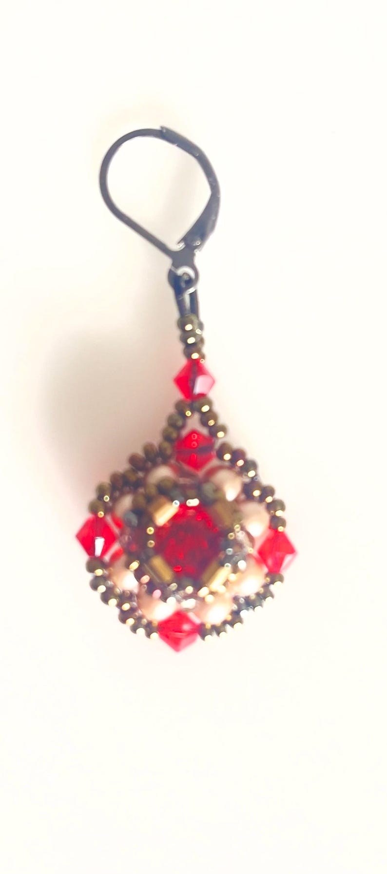 Może przedstawiać: Ręcznie robiony kolczyk w kształcie łzy. Posiada centralny czerwony kamień szlachetny, otoczony mniejszymi czerwonymi i złotymi koralikami. Kolczyk ma czarne zapięcie.