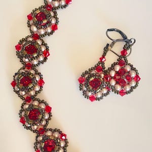 Może przedstawiać: Zestaw biżuterii z koralik&oacute;w składający się z bransoletki i kolczyk&oacute;w. Bransoletka i kolczyki mają powtarzający się kwiatowy wz&oacute;r z czerwonymi kwadratowymi kryształami, małymi białymi koralikami i metalowymi akcentami w kolorze brązu. Kolczyki mają zapięcie dźwigniowe.