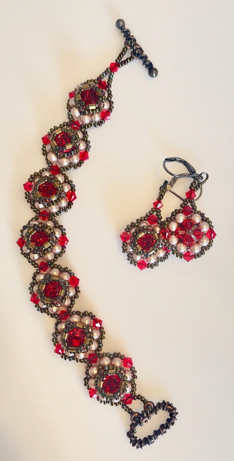 Może przedstawiać: Zestaw biżuterii z koralik&oacute;w składający się z bransoletki i kolczyk&oacute;w. Bransoletka i kolczyki mają powtarzający się kwiatowy wz&oacute;r z czerwonymi kwadratowymi kryształami, małymi białymi koralikami i metalowymi akcentami w kolorze brązu. Kolczyki mają zapięcie dźwigniowe.