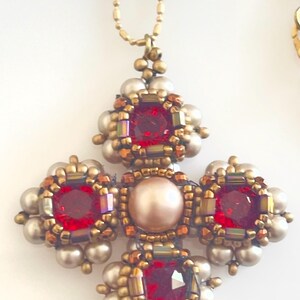 May include: A handcrafted pendant necklace featuring a cross design. The pendant has four red square gemstones, a central pearl, and is surrounded by gold-toned beads and smaller pearls. The necklace chain is gold-colored.
