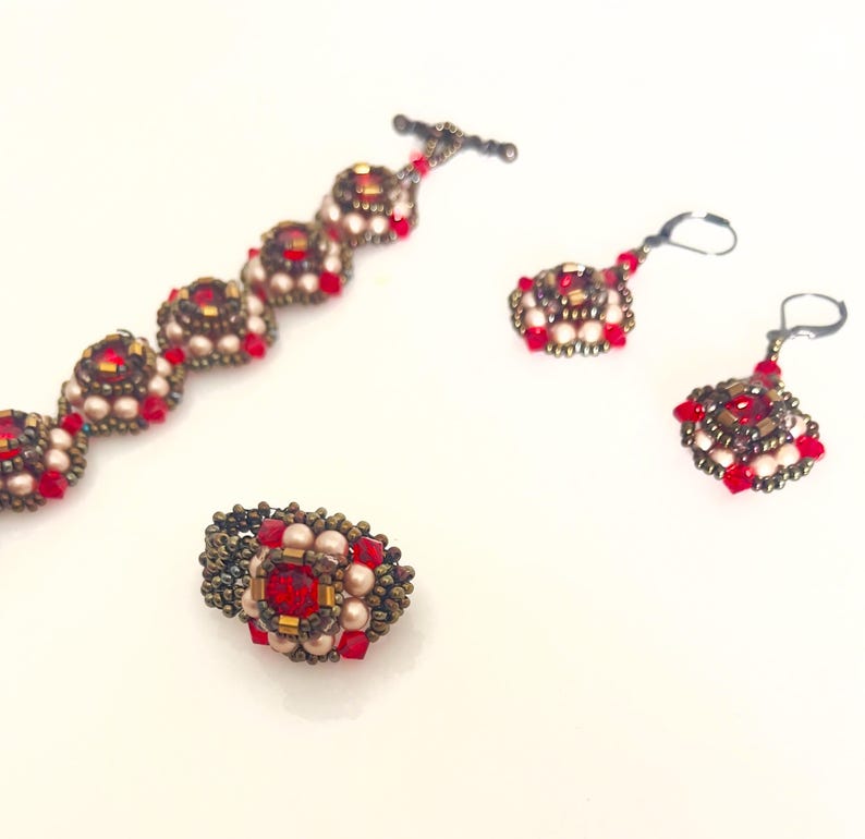 Może przedstawiać: Zestaw biżuterii składający się z bransoletki, pierścionka i kolczyk&oacute;w. Elementy zdobione są czerwonymi i perłowymi koralikami oraz centralnym czerwonym kamieniem. Metal ma brązowe wykończenie, tworząc estetykę vintage.