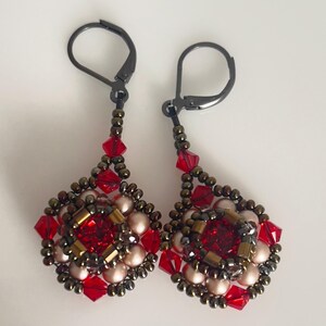 Może przedstawiać: Para ręcznie robionych kolczyk&oacute;w wiszących. Każdy kolczyk ma centralny czerwony kryształ otoczony czerwonymi, złotymi i perłowymi koralikami. Kolczyki mają czarne zapięcia typu leverback i mają kształt łzy.