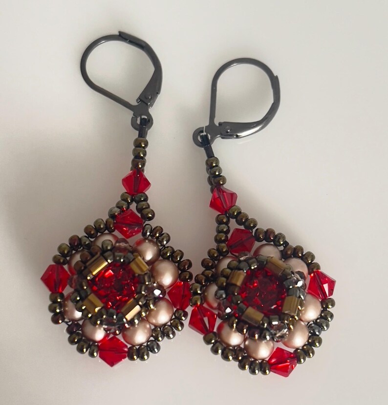 Może przedstawiać: Para ręcznie robionych kolczyk&oacute;w wiszących. Każdy kolczyk ma centralny czerwony kryształ otoczony czerwonymi, złotymi i perłowymi koralikami. Kolczyki mają czarne zapięcia typu leverback i mają kształt łzy.