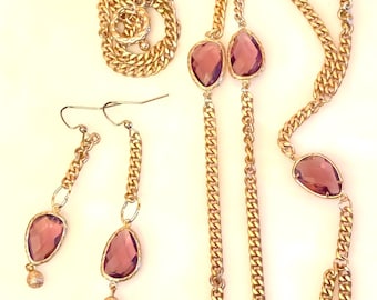 May include: A gold-tone jewelry set consisting of a long chain necklace and a pair of drop earrings. The necklace features multiple teardrop-shaped purple stones set in gold frames along a curb chain. The earrings match with similar stones.