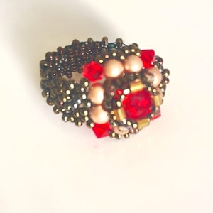 Może przedstawiać: Pierścionek z koralik&oacute;w z centralnym skupiskiem czerwonych i brzoskwiniowych koralik&oacute;w, otoczonych mniejszymi złotymi i brązowymi koralikami. Opaska pierścionka wykonana jest z ciasno tkanych brązowych koralik&oacute;w, tworząc teksturowany wygląd. Pierścionek jest na białym tle.