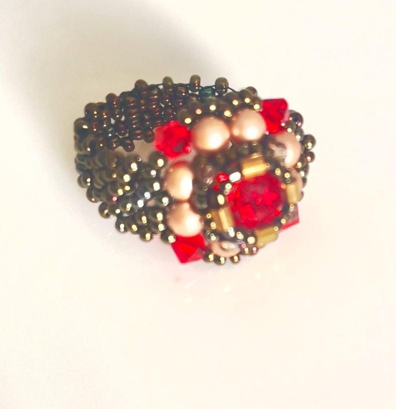 Może przedstawiać: Pierścionek z koralik&oacute;w z centralnym skupiskiem czerwonych i brzoskwiniowych koralik&oacute;w, otoczonych mniejszymi złotymi i brązowymi koralikami. Opaska pierścionka wykonana jest z ciasno tkanych brązowych koralik&oacute;w, tworząc teksturowany wygląd. Pierścionek jest na białym tle.