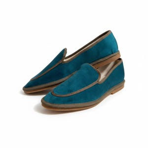 May include: A pair of teal velvet loafers featuring a pointed toe silhouette, a low heel, and a contrasting brown fabric trim along the collar and the vamp. The interior lining is a light beige color, and the outsole is a flat, light brown material.