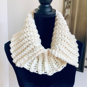 May include: A white crocheted cowl with a ruffled edge. The cowl is displayed on a black mannequin.