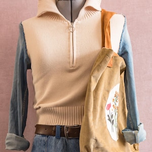 Op de afbeelding: Een crèmekleurige, geribbelde gebreide trui met kwartrits en lange mouwen van spijkerstof, gecombineerd met een blauwe spijkerrok met een bloemenapplicatie. Een beige corduroy tas met een wit ovaal en geborduurde wilde bloemen.