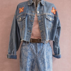 Op de afbeelding: Lichtblauwe spijkerjas met oranje bloemenpatches op de schouders, gecombineerd met een bijpassende spijkerrok met bloemenpatroon. Een gestreept shirt is zichtbaar onder de jas en een bruine riem accentueert de taille.