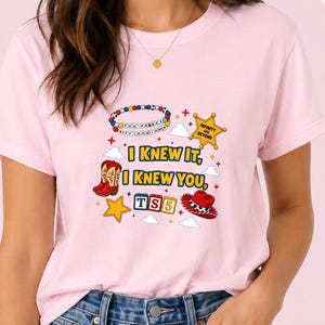 May include: A light pink t-shirt featuring a graphic print with the text I KNEW IT, I KNEW YOU, TS5, and INFINITY AND BEYOND. The design includes illustrations of a beaded bracelet, a sheriff star, cowboy boots, a cowboy hat, and stars.