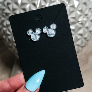 May include: A pair of stud earrings shaped like mouse heads with a geometric, multifaceted silver and white pattern. The earrings are attached to a rectangular black display card with a small hole at the top for hanging, showcasing a clean design.